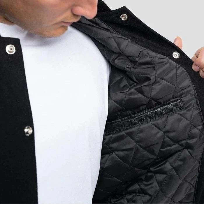 Black varsity jacket with leather sleeves and orange accents worn by a person on a light gray background